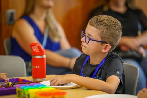 A young boy wearing glasses and a blue lanyard sits at a table with a red water bottle and colorful tubs of playdough in front of him.